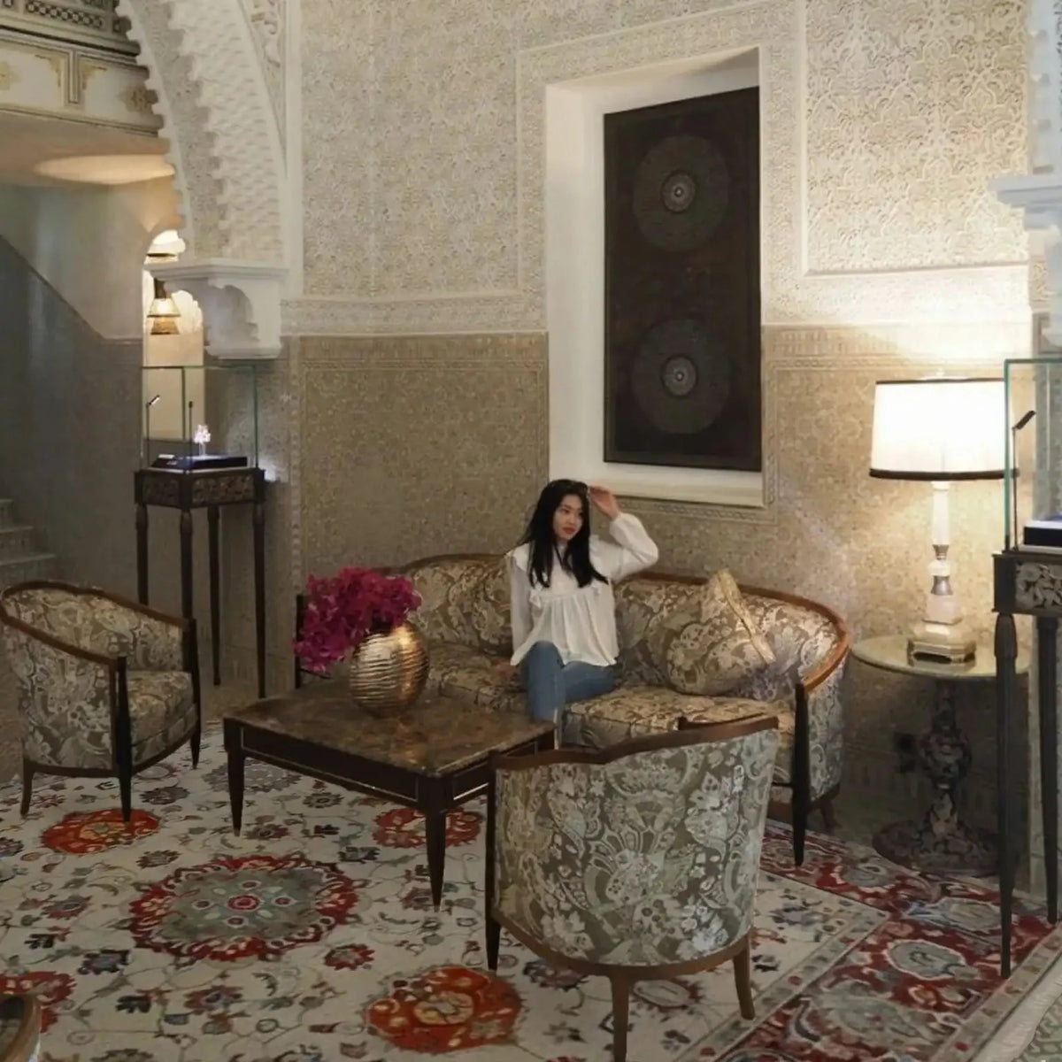 Woman sitting on a couch in an ornate room with intricate floral persian rug in red and beige color, wall patterns and decorative furniture.