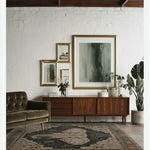 A beautifully styled living room featuring a large Elephant Whisper hand-tufted wool rug. The area rug, perfect for living rooms, showcases an intricate design with a central elephant motif. It sits on a wooden floor, complementing the mid-century modern furniture, including a plush green sofa and a sleek wooden sideboard adorned with framed art and greenery.