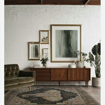 A beautifully styled living room featuring a large Elephant Whisper hand-tufted wool rug. The area rug, perfect for living rooms, showcases an intricate design with a central elephant motif. It sits on a wooden floor, complementing the mid-century modern furniture, including a plush green sofa and a sleek wooden sideboard adorned with framed art and greenery.