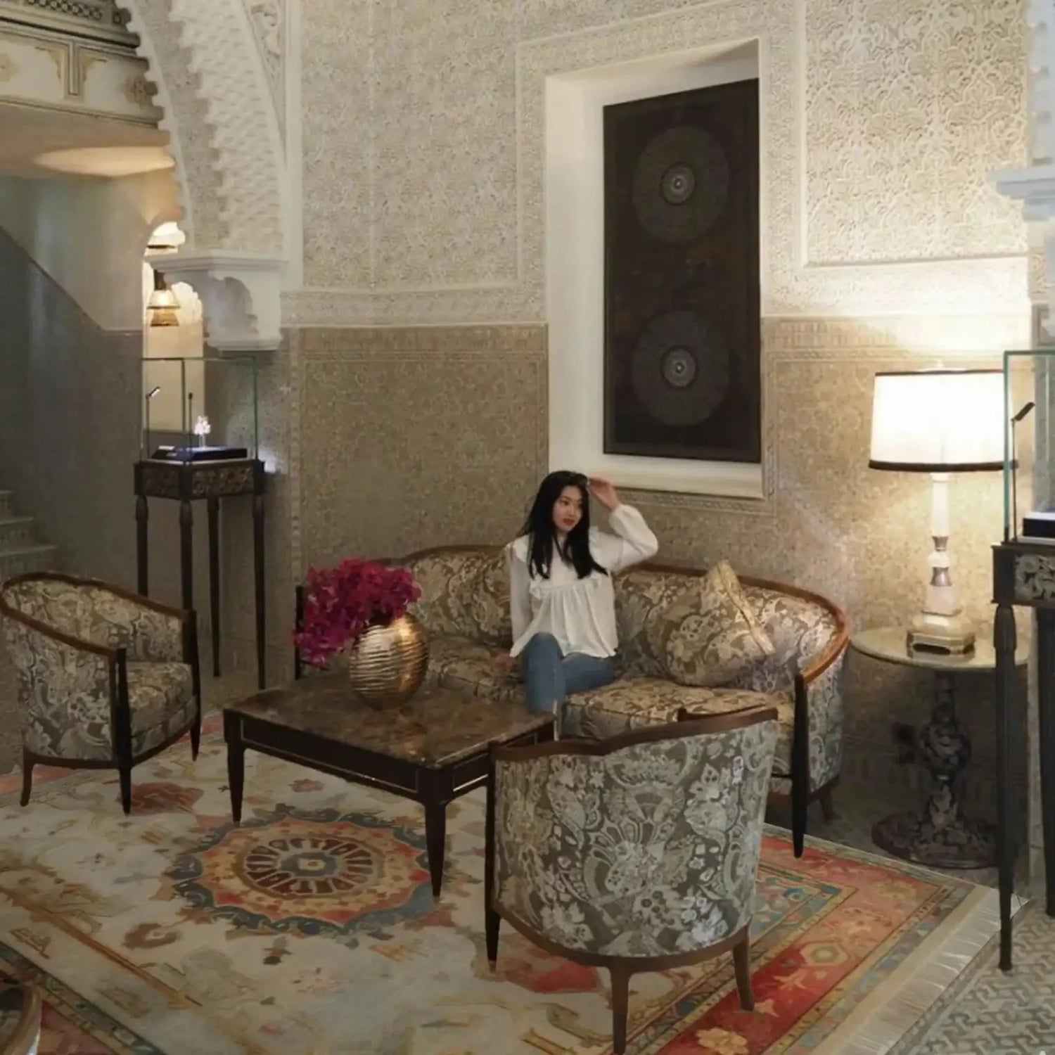 Woman sitting on a couch in an ornate room with intricate beige handmade rug, wall patterns and decorative furniture.