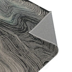 Folded corner showing charcoal surface with flowing grey wave line pattern and grey fabric backing with overlock stitching on the Ethereal Archway Fluid Pathways Rug