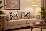 A beige upholstered sofa in a warm, styled living room setting, featuring two decorative throw pillows with a blue, orange, and cream horse pattern. A wooden coffee table, a floor lamp, and potted plants are visible in the background.