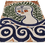 Flat view of a hand-tufted rug depicting a serene face with fern-frond hair above navy swirls on cream wool