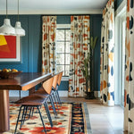 Floral Extravaganza Wool Hand Knotted Area Rug in a dining room setting. The vibrant floral rug complements the modern decor with its rich colors and intricate patterns, perfect for living room and dining room spaces. This Persian-inspired wool rug enhances the wooden dining table and chairs, framed by abstract-patterned curtains and contemporary wall art.