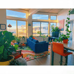Modern living room with blue sofa, orange chair on a rug and large windows.