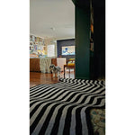 A stylish living room featuring the Flowing Illusion Black and Gray Wool Runner Rug with bold black and gray stripes, perfect as an area rug for contemporary spaces. This vintage rug elegantly complements the decor of a cozy living room, enhancing the charm of wooden flooring and eclectic wall art. Ideal for use as a hallway runner rug or in a large living room setting.