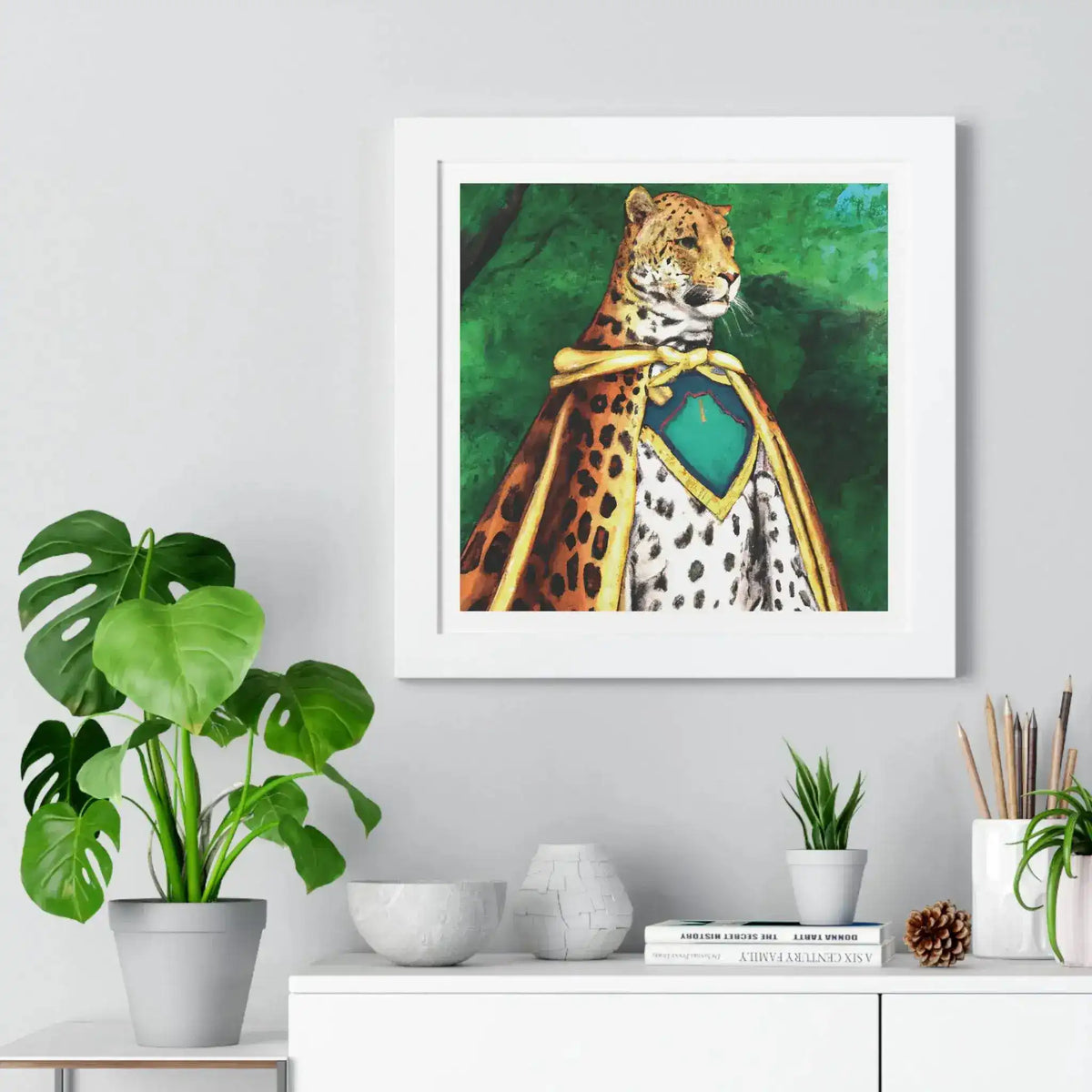 Framed horizontal poster in white frame, leopard in gold-trimmed cape on green background, above white sideboard with plant