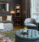 A stylish living room featuring the Garden of Courage Hand Tufted Rug in a night theme. This large area rug showcases a vintage design with a floral pattern, adding elegance to the space. The wool rug complements the sophisticated decor, including a tufted ottoman and plush seating, perfect for enhancing any living room area.