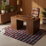 Wooden desk with leather chair in a room with plants, maroon rug, and a lamp.
