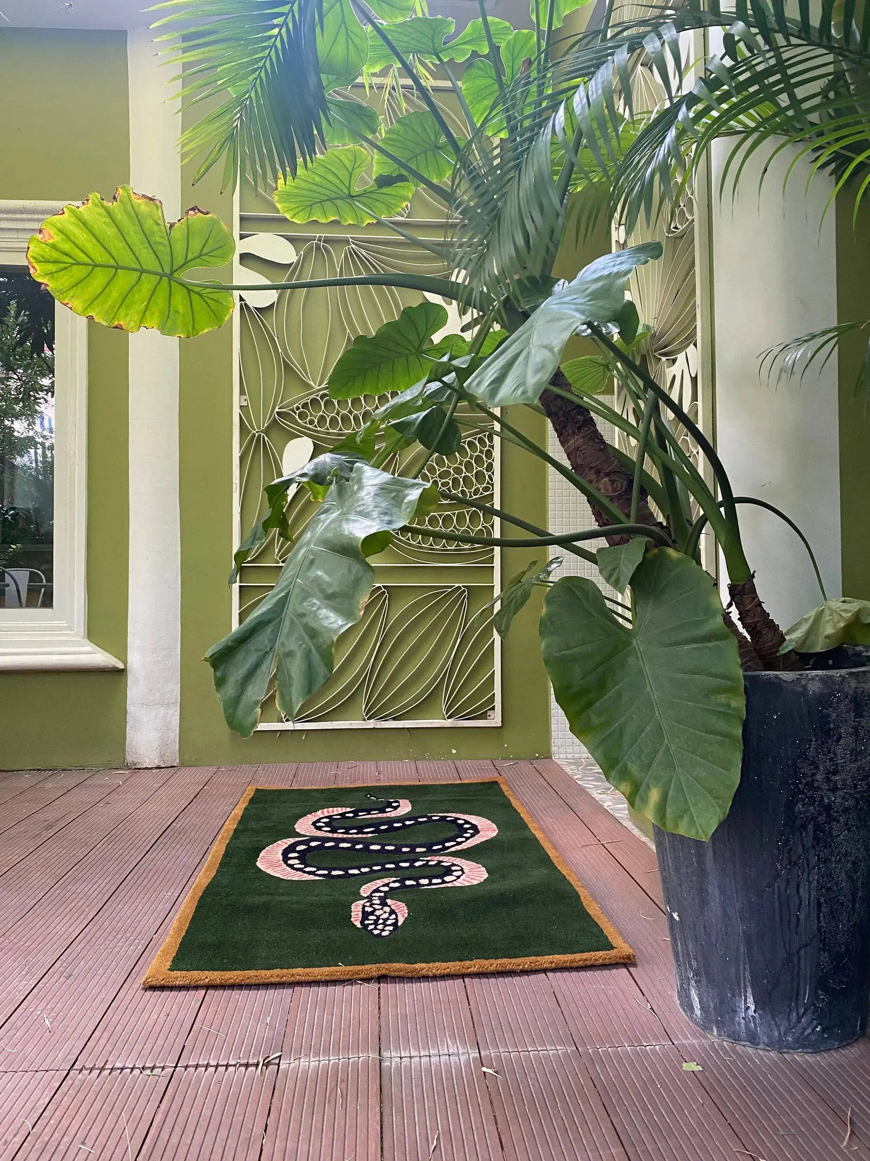 The Green and Black Snake Hand Tufted Wool Area Rug is elegantly displayed on a wooden patio surrounded by lush greenery. This vintage rug, perfect for a dining room setting, features a striking snake design on a deep green background, making it an ideal choice for a dining table rug or living room rugs 8x10. The wool rug's intricate design adds a touch of sophistication to any living room decor.