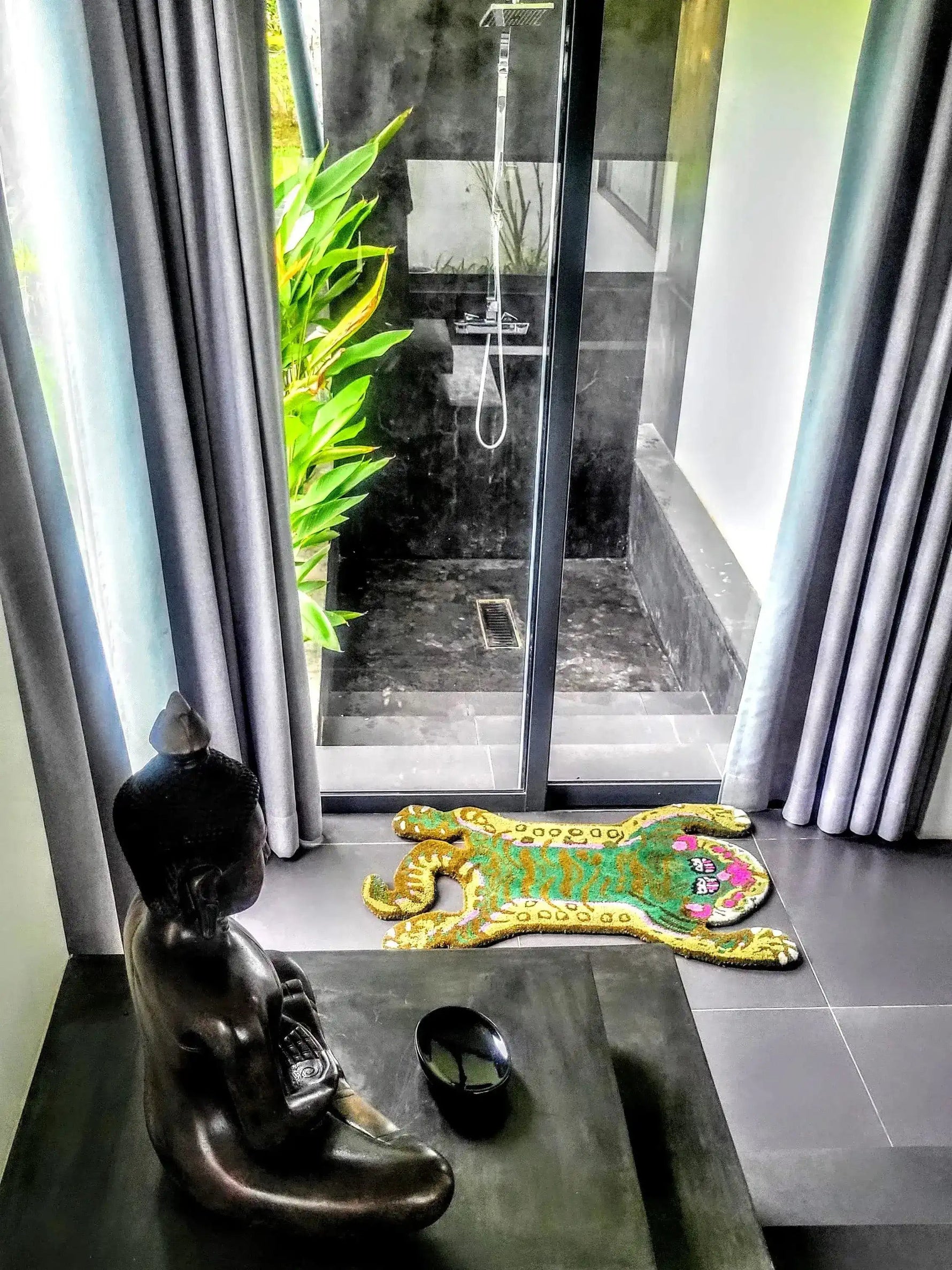 A vibrant Green Asian Tiger Wool Rug placed next to a serene statue in a modern room with glass doors leading to an outdoor shower space. This abstract vintage rug adds a unique touch to any dining room, office, or formal dining area, showcasing its versatile use as a dining room carpet.