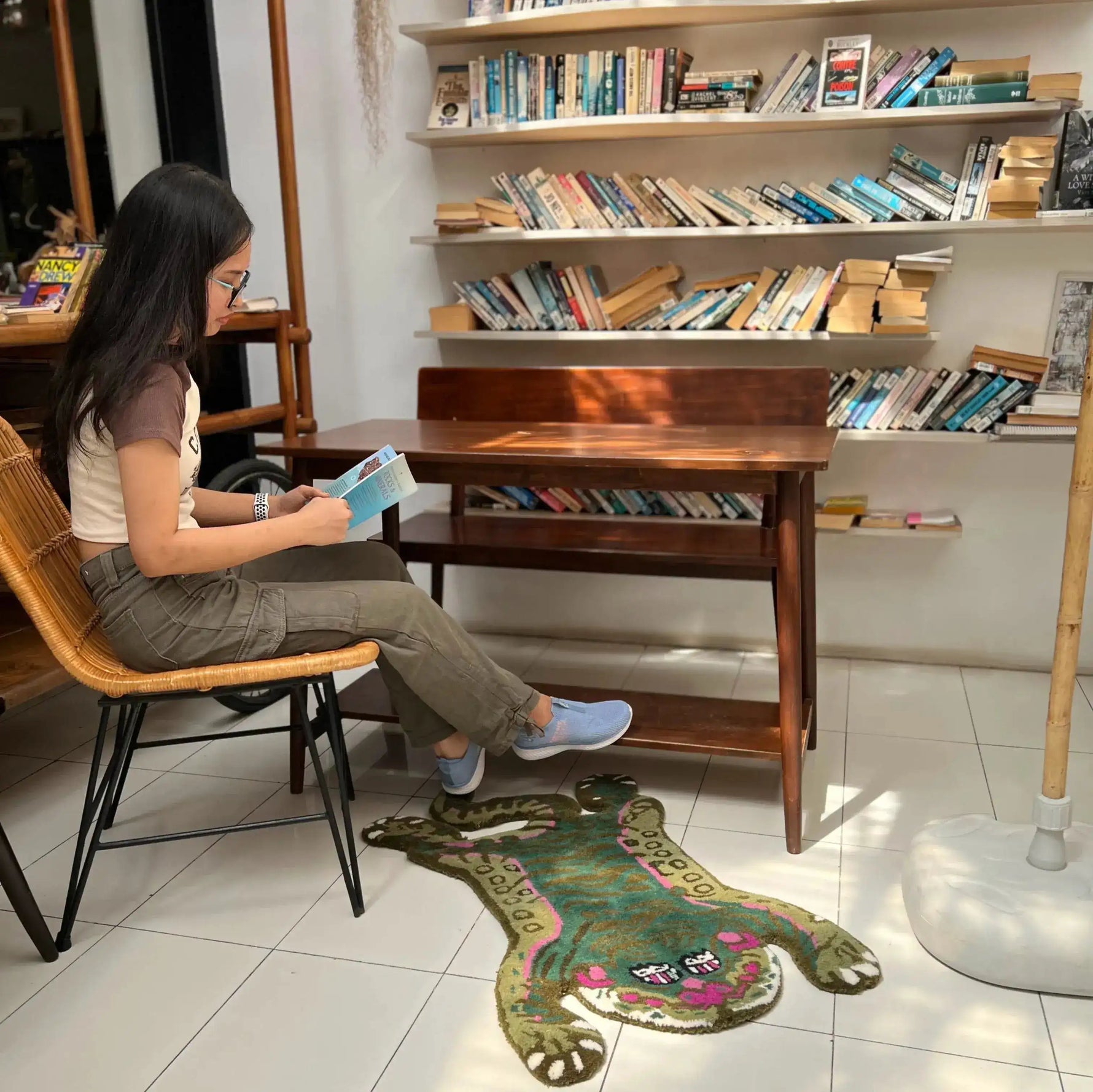A cozy reading nook featuring a unique Green Asian Tiger Wool Rug. The area rug adds a touch of whimsy to the space, ideal for a vintage rug feel in a dining room or office. A woman is seated on a wicker chair, absorbed in a book, highlighting how this wool rug can complement formal dining room rugs or dining room carpets.