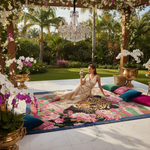 Woman in a gold dress sitting on a colorful rug with floral decorations and a chandelier in a luxurious garden setting.
