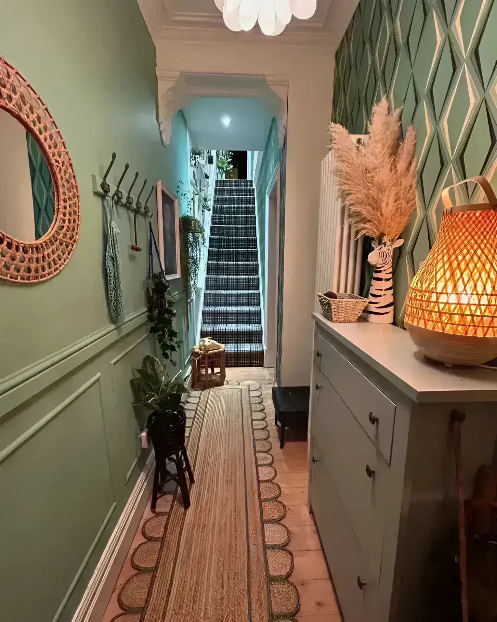 A charming hallway featuring a handmade olive green scalloped border jute rug, perfect as an area rug or hallway runner rug. The rug complements the elegant decor, including a wicker mirror, plants, and a stylish cabinet with a warm lamp. Ideal for adding a cozy touch to any living room or kitchen space.