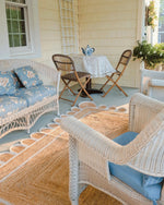 Charming outdoor seating area featuring a handmade white scalloped jute area rug, perfect for a dining room or living room. The natural texture of the rug complements the wicker furniture, creating a cozy and inviting space ideal for relaxing or entertaining guests.