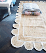 Close-up view of a handmade white scalloped jute rug laid on a dark hardwood floor, perfect for dining room or living room areas. The natural texture and elegant design make it an ideal area rug choice for adding warmth and style to any living space.