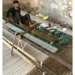 A skilled artisan handweaving a green cotton rug featuring an intricate palm tree design. The rustic setting highlights the craftsmanship involved in creating this boho area rug, perfect for a dining room or as an 8x10 living room centerpiece.