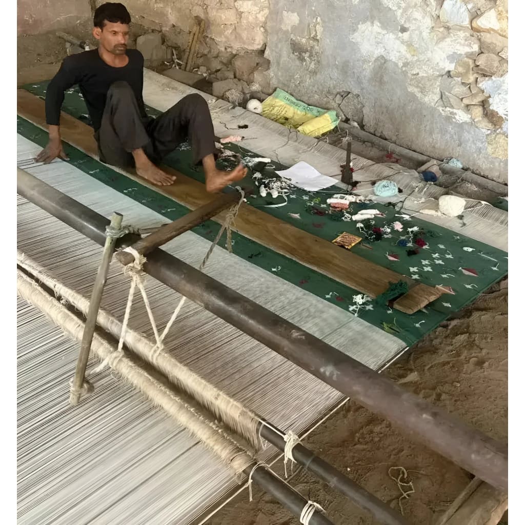 A skilled artisan handweaving a green cotton rug featuring an intricate palm tree design. The rustic setting highlights the craftsmanship involved in creating this boho area rug, perfect for a dining room or as an 8x10 living room centerpiece.