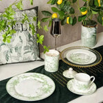 White ceramic dinnerware set with green lily of the valley botanical print, styled on dark green velvet table runner with lemon tree backdrop