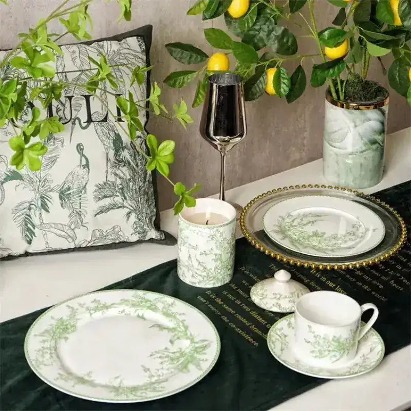 White ceramic dinnerware set with green lily of the valley botanical print, styled on dark green velvet table runner with lemon tree backdrop