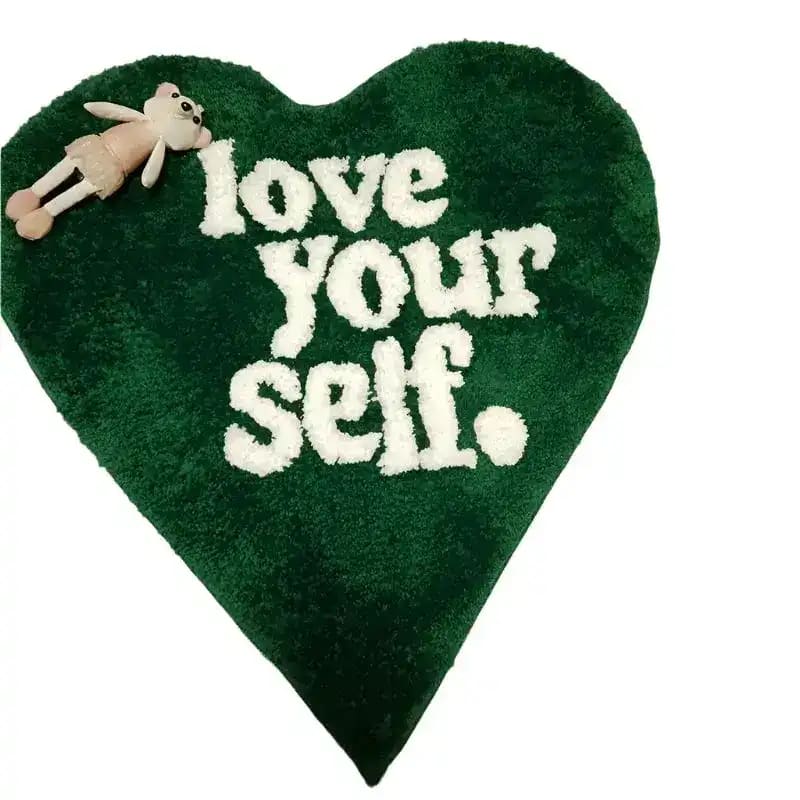 Heart-shaped area rug in a deep green color with the words 'Love Your Self' in bold white text. A small plush toy is placed on the corner, adding a playful touch. Perfect as an area rug for living rooms, this rug combines comfort with a positive message.
