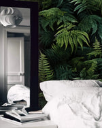 Lush green fern jungle wallpaper behind white bedding with tall floor mirror and books