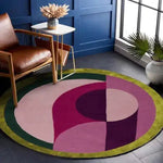 A modern Maia Golden Pink Round Hand Tufted Wool Rug displayed in a stylish living room. The round area rug features an abstract design in shades of pink and purple with a contrasting green border, perfect for adding a contemporary touch to a living space. Ideal as a large area rug for living room settings, this 8x10 wool rug complements various decor styles.