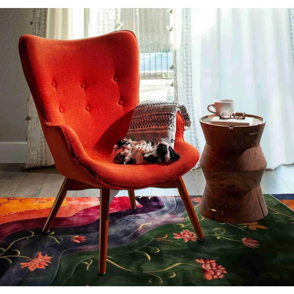 A vibrant, hand-tufted area rug featuring an abstract floral design in rich colors graces the floor of a stylish dining room. The wool rug adds a touch of elegance and warmth, complementing a striking orange chair and a wooden side table, perfect for both dining room and office settings.