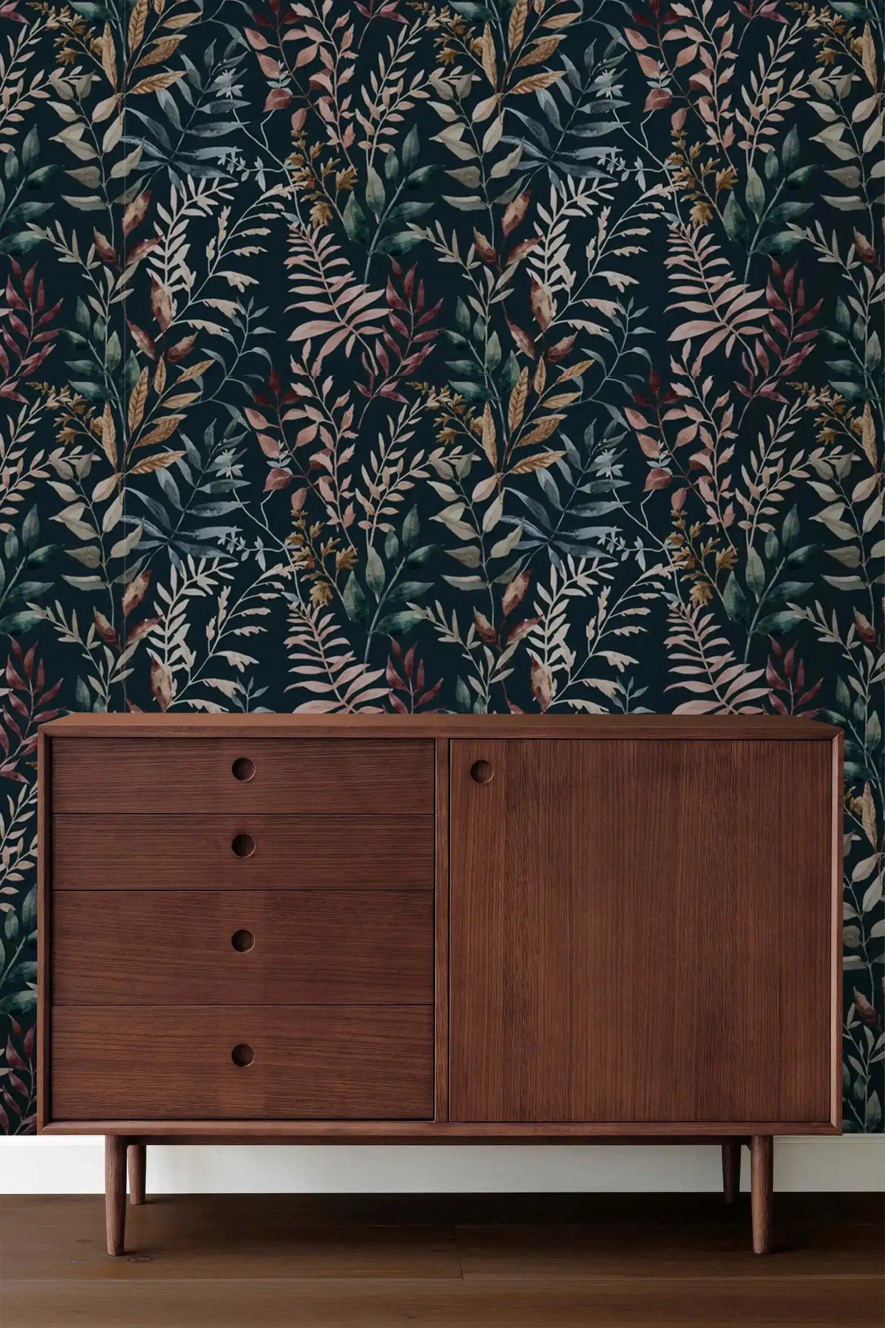 Midnight botanical wallpaper with mixed-color leaves behind mid-century dark walnut sideboard with drawers and cabinet.