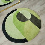 A stylish and modern Mini Maia geometric round hand-tufted wool rug in shades of green and black, placed on a light wooden floor. This area rug serves as an elegant addition to large living rooms or offices, enhancing the space with its abstract design and cozy texture.