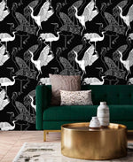 Monochrome crane wallpaper with white line-drawn cranes on black behind an emerald velvet sofa and gold table