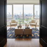 A modern living room interior with two cream armchairs facing two low light-wood coffee tables on a dark patterned area rug. Large floor-to-ceiling windows overlook a city skyline and green park area. The room is framed by open dark wood doors.