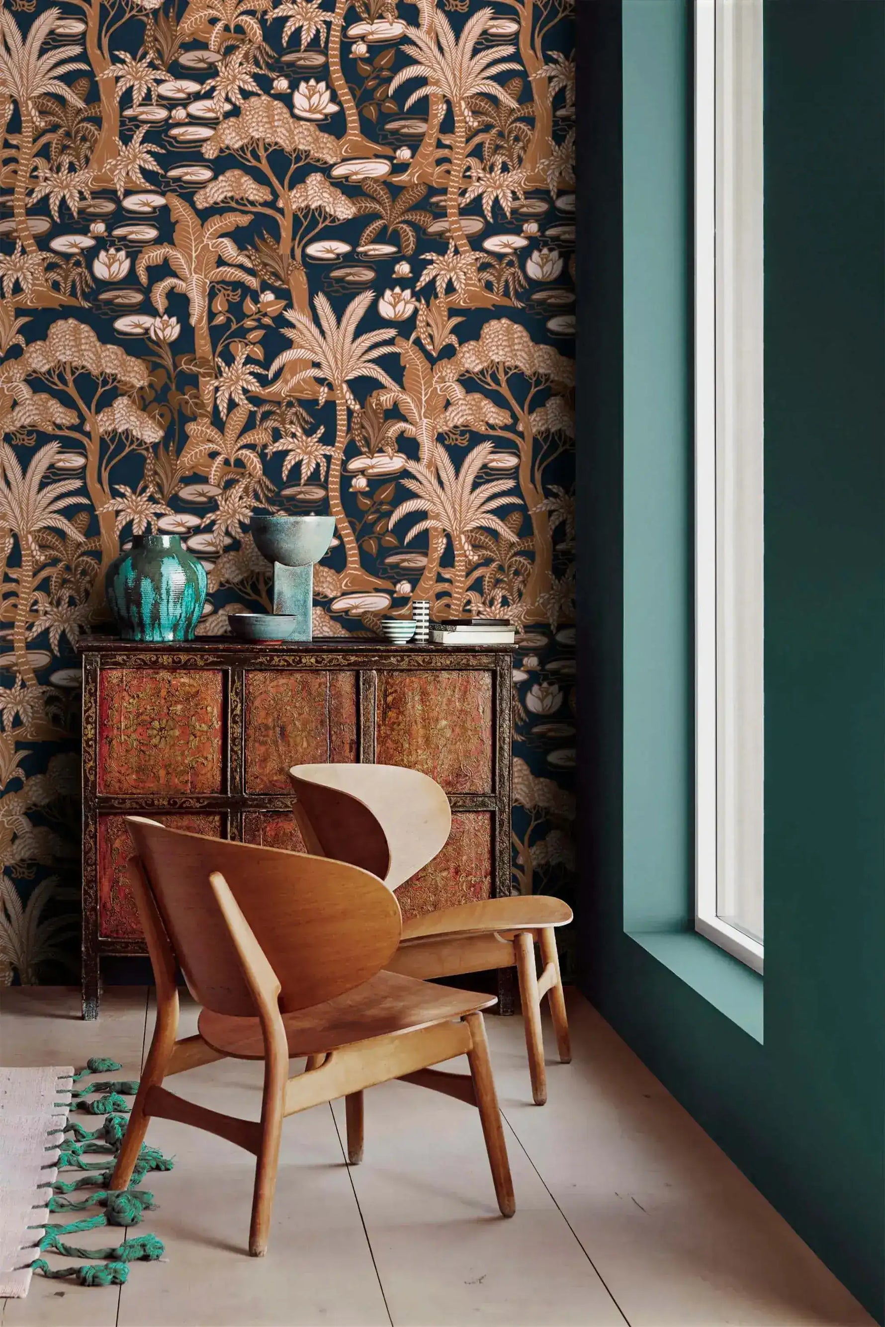 Navy copper palm wallpaper beside a teal wall, with antique cabinet and curved wood chairs