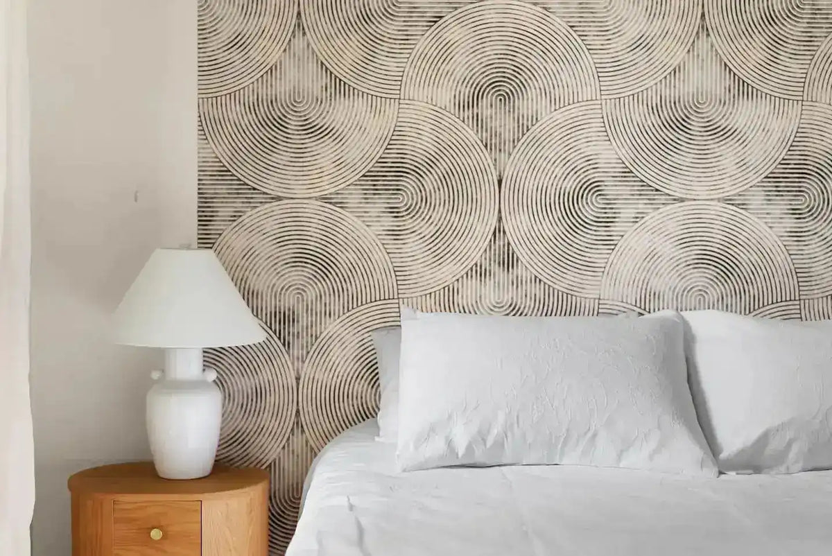 Close-up of beige concentric-circle geometric wallpaper beside a bed with a white lamp and oak nightstand