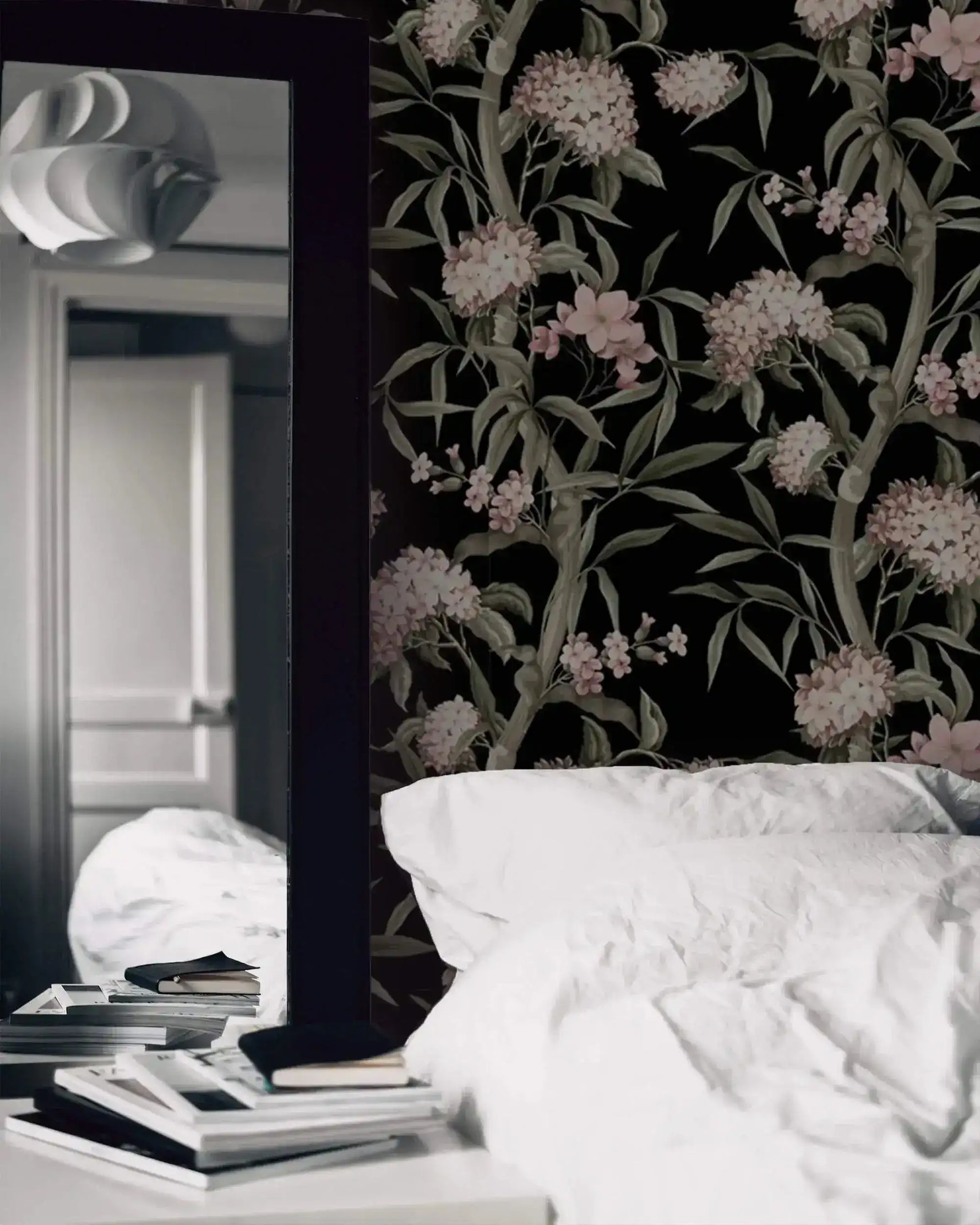 Blushing blossoms wallpaper beside dark-framed mirror, white bedding and stacked books on nightstand in bedroom