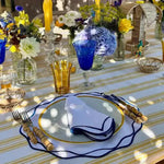 Ocean ripple placemat with navy wavy edge, gold-rimmed charger, blue folded napkin, outdoor florals in background