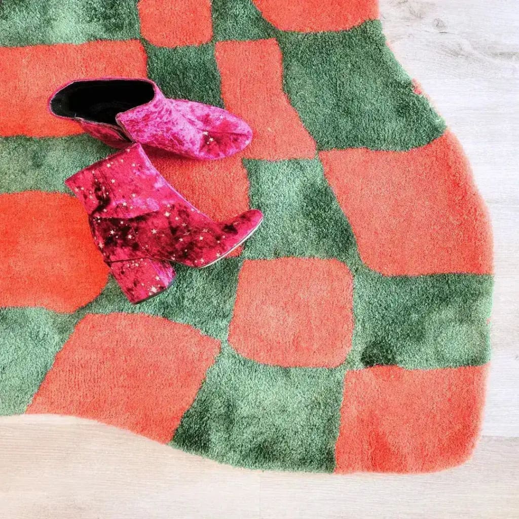 Vibrant orange and green wavy checker hand-tufted wool area rug, perfect for adding a pop of color to your living room or as a unique hallway runner rug. The abstract design of this large area rug complements various styles, making it a versatile choice for kitchen rugs or vintage rug enthusiasts. A pair of stylish pink boots rests on the rug, highlighting its plush texture.