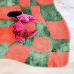 Vibrant orange and green wavy checker hand-tufted wool area rug, perfect for adding a pop of color to your living room or as a unique hallway runner rug. The abstract design of this large area rug complements various styles, making it a versatile choice for kitchen rugs or vintage rug enthusiasts. A pair of stylish pink boots rests on the rug, highlighting its plush texture.