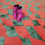 A vibrant orange and green wavy checker pattern on a hand-tufted wool area rug, perfect for enhancing your living room, kitchen, or hallway. This large area rug features a unique abstract design with a pair of purple velvet boots resting on it, adding a touch of elegance to your space. Ideal as a living room rug in an 8x10 size or as a vintage rug for any room.