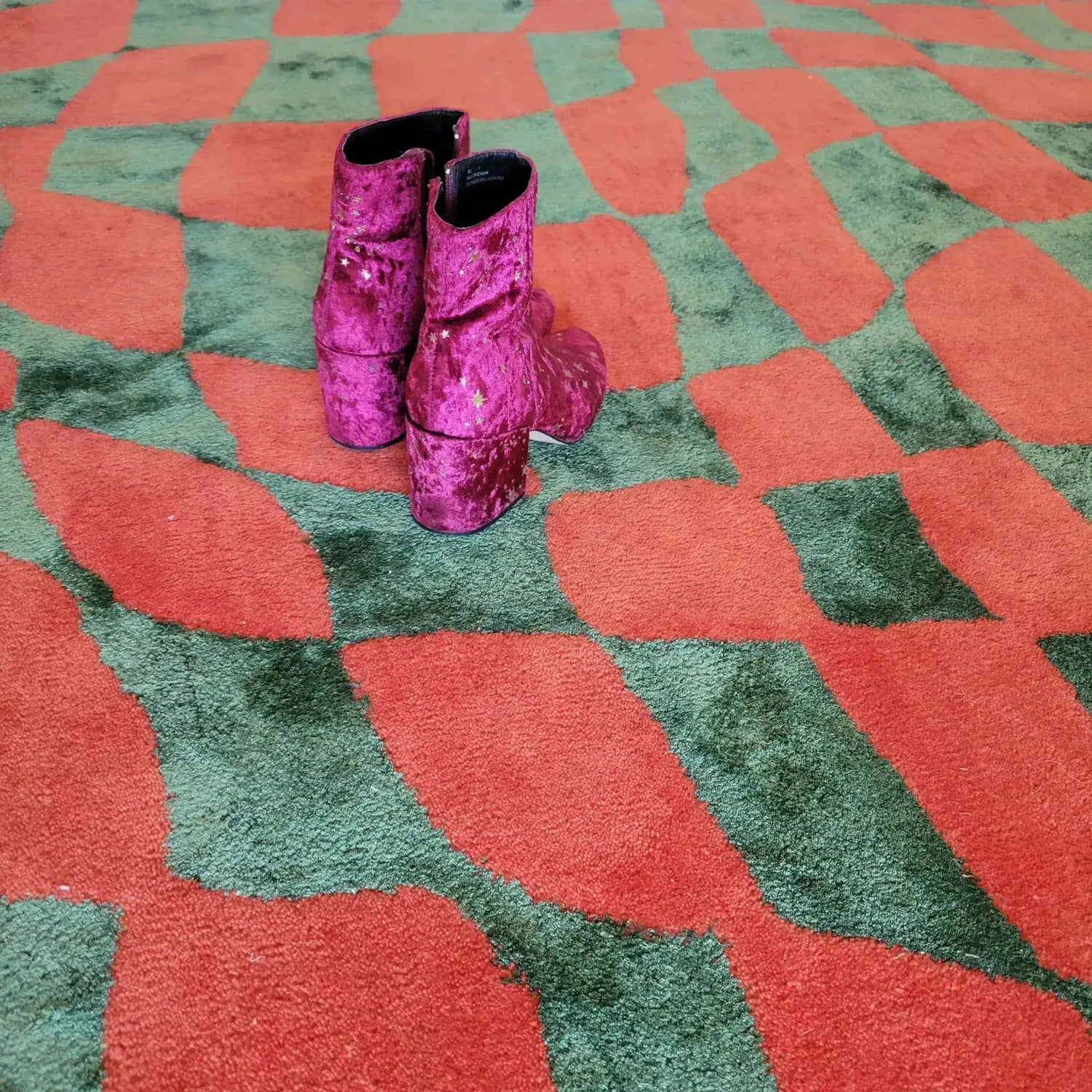 A vibrant orange and green wavy checker pattern on a hand-tufted wool area rug, perfect for enhancing your living room, kitchen, or hallway. This large area rug features a unique abstract design with a pair of purple velvet boots resting on it, adding a touch of elegance to your space. Ideal as a living room rug in an 8x10 size or as a vintage rug for any room.