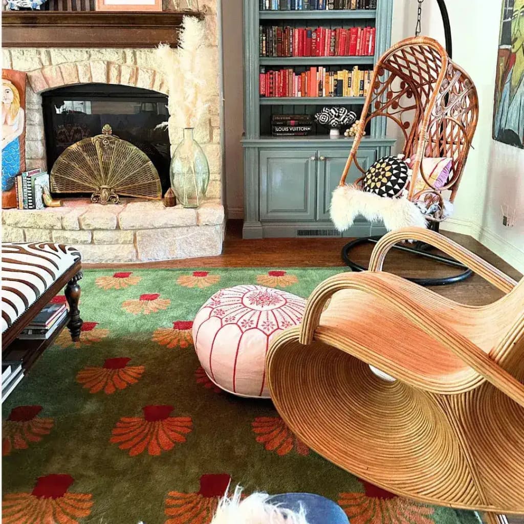 Cozy office space featuring a large area rug with vibrant orange palm tree patterns on a green tufted wool surface. The wool rug adds warmth and style to the room, complementing the eclectic decor with a hanging chair, pouf, and stone fireplace.