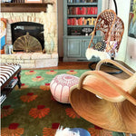 Cozy office space featuring a large area rug with vibrant orange palm tree patterns on a green tufted wool surface. The wool rug adds warmth and style to the room, complementing the eclectic decor with a hanging chair, pouf, and stone fireplace.