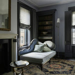 Decorative chaise lounge in a stylish room with dark floral traditional rug, bookshelf and window.