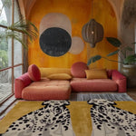 A vibrant living room featuring a contemporary wool rug with an abstract dog design in mustard, complementing a large, cozy pink sectional sofa. The area rug adds a modern touch to the large space, enhanced by the colorful wall art and a woven pendant light.