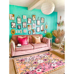 A vibrant living room featuring a pastel paradise hand tufted area rug with a floral design. The rug, perfect for living room area rugs, adds a vintage touch with its colorful wool texture. The room is arranged with a pink sofa adorned with cushions, under a gallery wall of floral art, creating a cozy and stylish atmosphere.