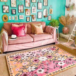 A vibrant floral area rug in a living room setting, featuring a hand-tufted wool design with pink and orange hues. The rug is situated in front of a cozy pink sofa adorned with colorful cushions. The walls are decorated with framed botanical art, creating a vintage and inviting atmosphere. Perfect for adding a touch of elegance to any living room area.