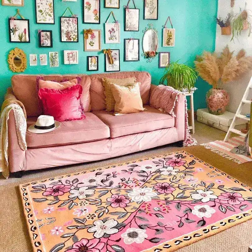 A vibrant floral area rug in a living room setting, featuring a hand-tufted wool design with pink and orange hues. The rug is situated in front of a cozy pink sofa adorned with colorful cushions. The walls are decorated with framed botanical art, creating a vintage and inviting atmosphere. Perfect for adding a touch of elegance to any living room area.