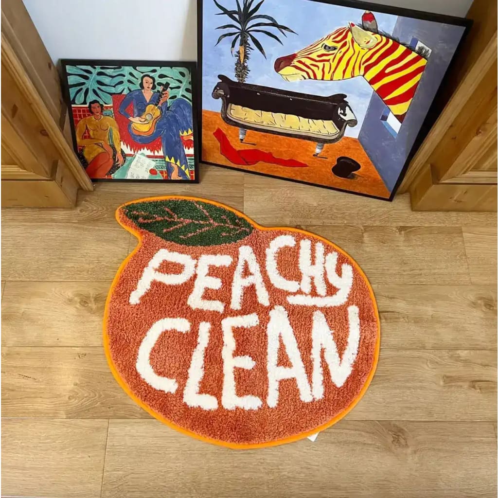 A vibrant Peachy Clean bathmat in the shape of a peach, featuring bold white text on an orange background, perfect as a runner rug for kitchens, hallways, or bathrooms. The cotton rug adds a playful touch to any space with its unique design, seen on a wooden floor next to colorful artwork.