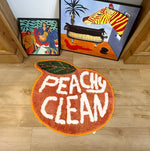 A vibrant Peachy Clean bathmat in the shape of a peach, featuring bold white text on an orange background, perfect as a runner rug for kitchens, hallways, or bathrooms. The cotton rug adds a playful touch to any space with its unique design, seen on a wooden floor next to colorful artwork.