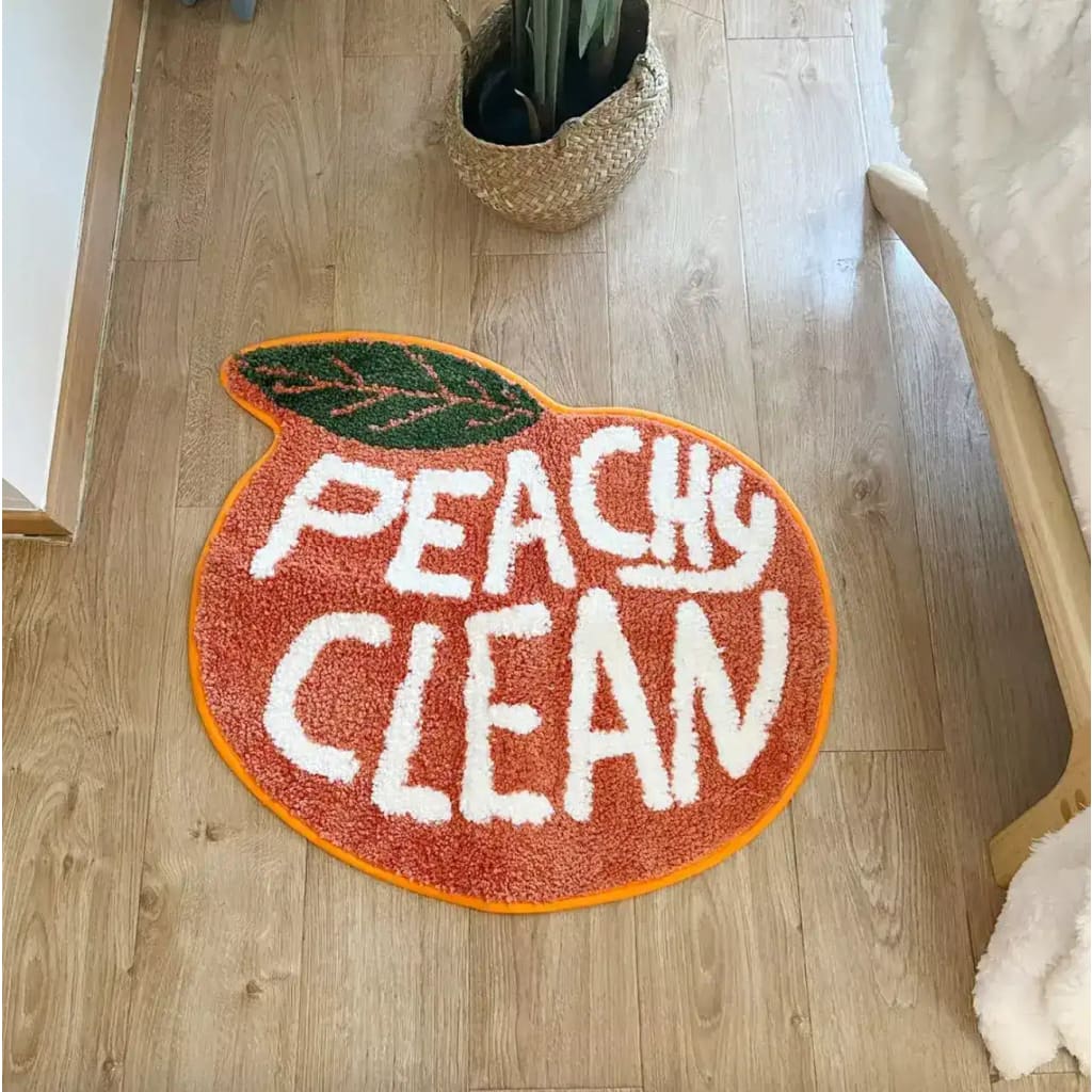 A vibrant peach-shaped bathmat with the words 'Peachy Clean' in bold white letters, perfect for adding a playful touch to your bathroom or kitchen. This cotton runner rug, ideal as a hallway runner rug or kitchen runner rug, is set against a warm wooden floor, offering a cozy and inviting atmosphere.